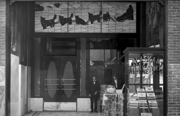 Historic black and white photo of the exterior of the Alaska Cigar Shop and Café featuring the lower half of the iconic Alaskan stained glass window.