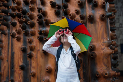 Girl wearing a rainbow umbrella hat looking through binoculars.