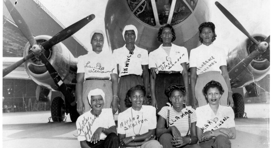 Eight black workers at Boeing during World War II pose in front of a large airplane with propellor engines on the left and right.