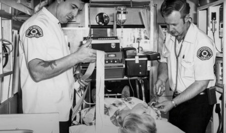 Two paramedics work on patient. One administers an IV while the other reviews an EKG rhythm strip.