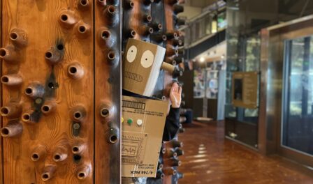 A cardboard robot waves at the camera while standing adjacent to Wawona wooden sculpture in MOHAI Grand Atrium.