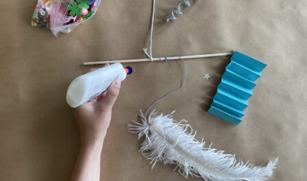 Single arm with hand holding glue, a wooden dowel, a white feather, and a clear plastic bag full of colorful knick knacks.