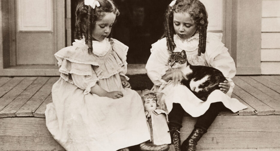 Two girls sitting with their cat and a doll on a porch in 1900.