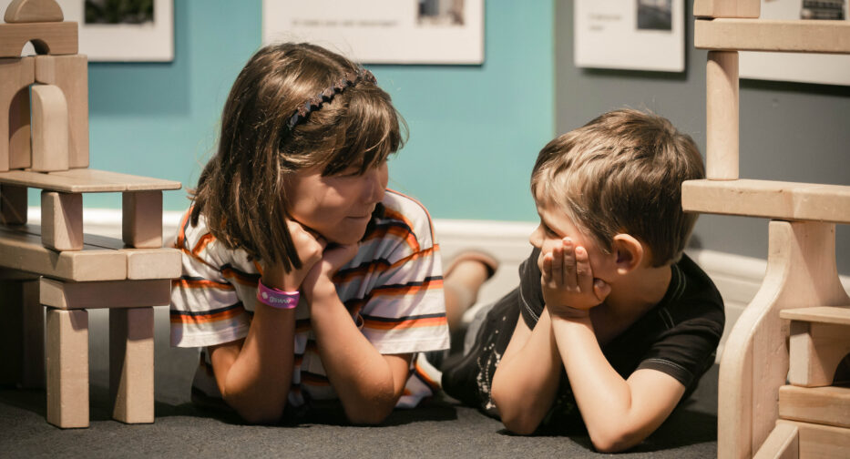 Two children look at each other while lying down playing with wooden blocks in the Kid-Struction Zone at MOHAI.