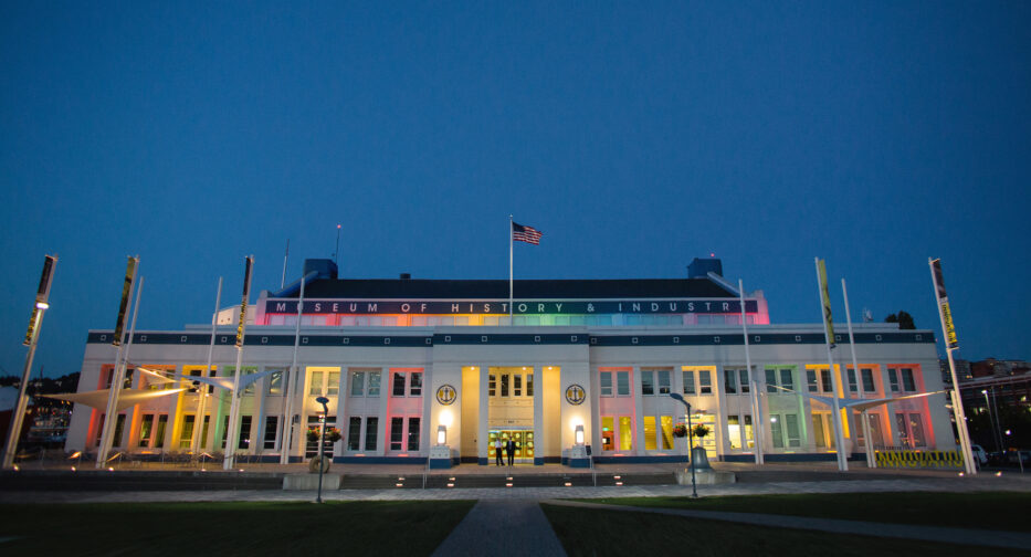 MOHAI exterior illuminated with rainbow lighting and seen at dusk below of a clear, blue sky.