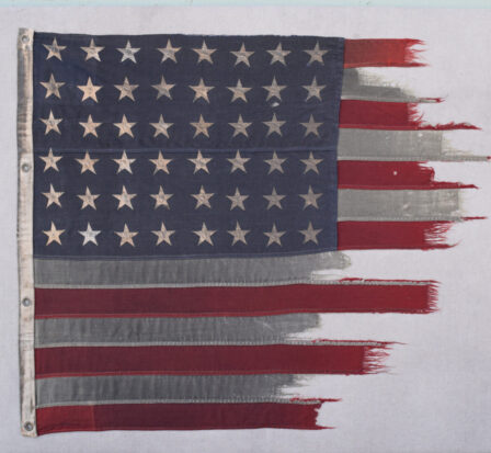 Tattered remains of an American flag flown on D-Day. The flag is red and white striped with a field of 48 white stars on a blue background in the upper left corner.