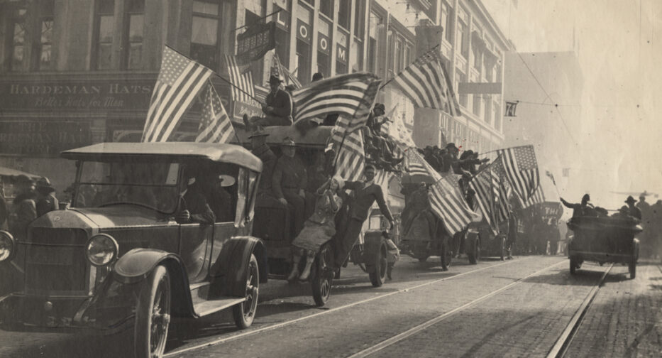 A parade of cars with riders waving flags in the Armistice Day parade.