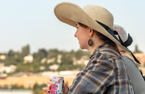 Woman wearing large sun hat drinking a grapefruit La Croix and facing to the left.