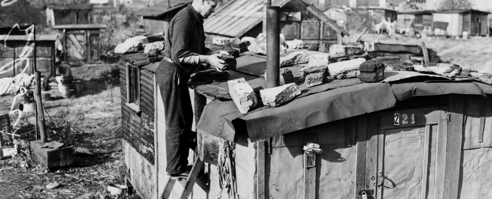 Black and white photo of Edwin Hill on ladder repairing the roof of his shack in Hooverville