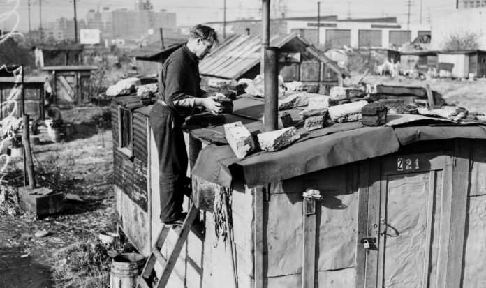 Black and white photo of Edwin Hill on ladder repairing the roof of his shack in Hooverville