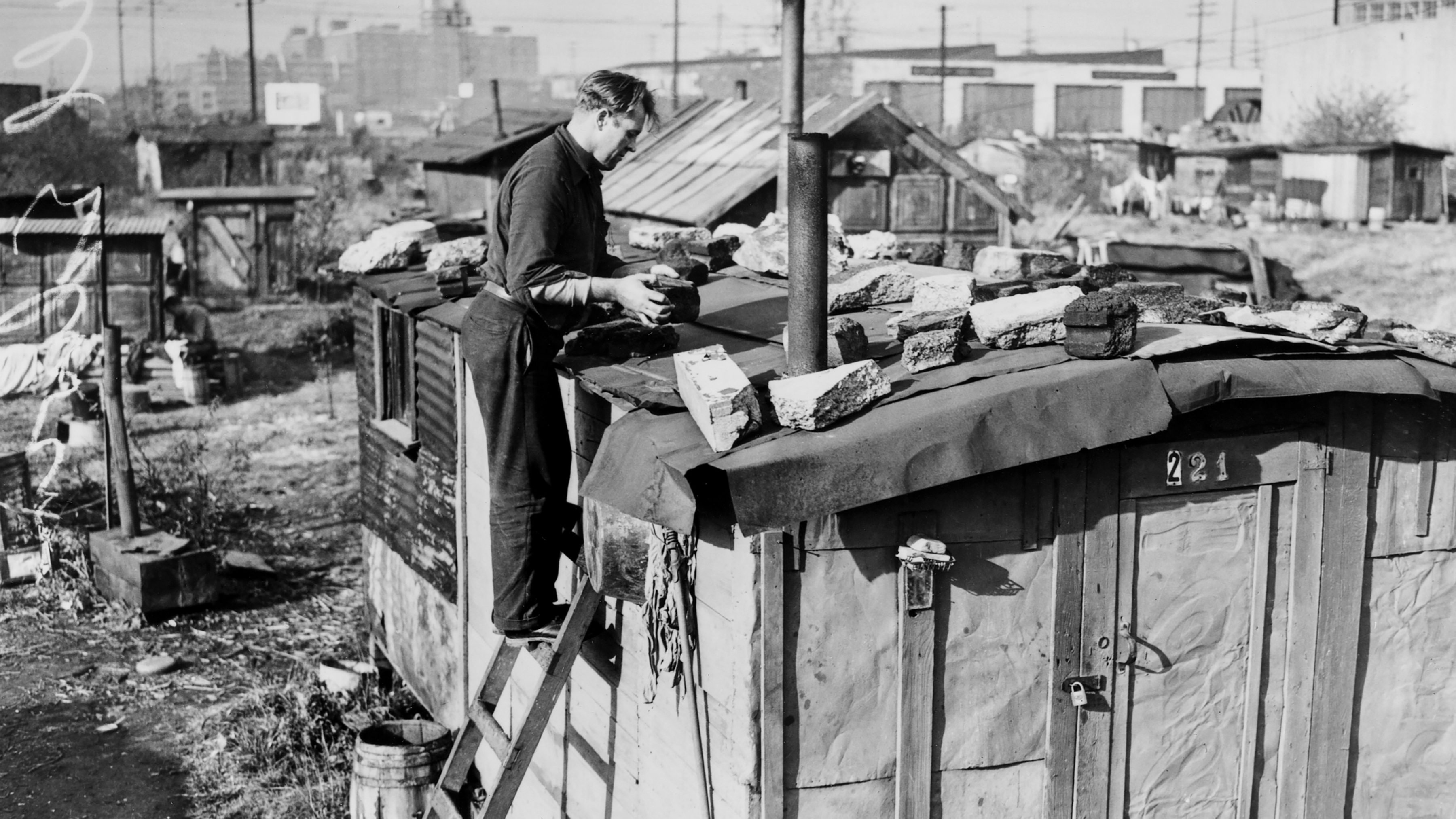 Black and white photo of Edwin Hill on ladder repairing the roof of his shack in Hooverville