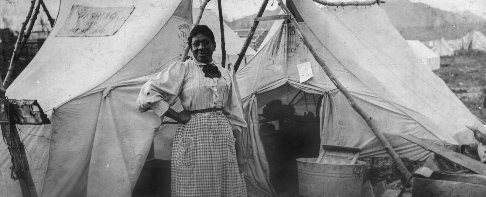 Black women standing in front of two white tents with washing board to her left.