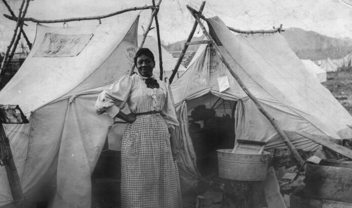 Black women standing in front of two white tents with washing board to her left.