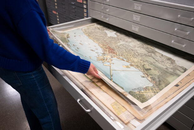 A person in a blue sweater stands before a variety of maps in MOHAI storage.