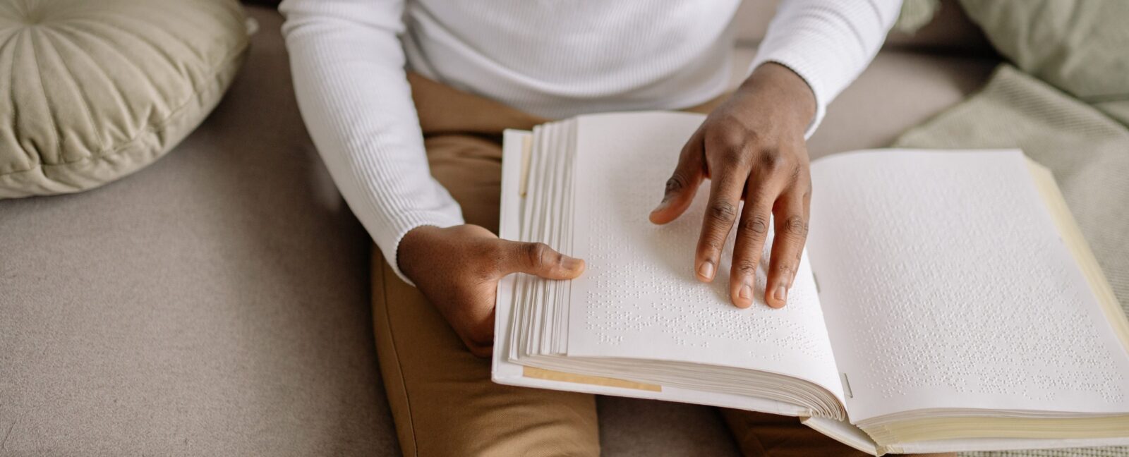 Person of color wearing white shirt and brown pants reading a book written in braille.