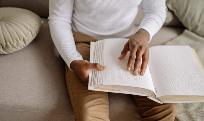 Person of color wearing white shirt and brown pants reading a book written in braille.