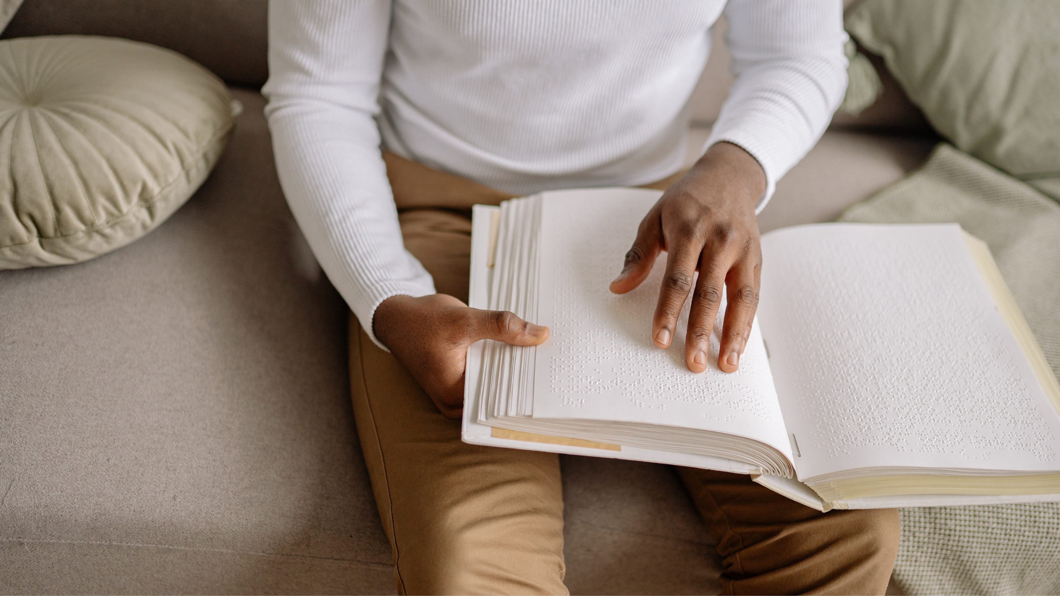 Person of color wearing white shirt and brown pants reading a book written in braille.