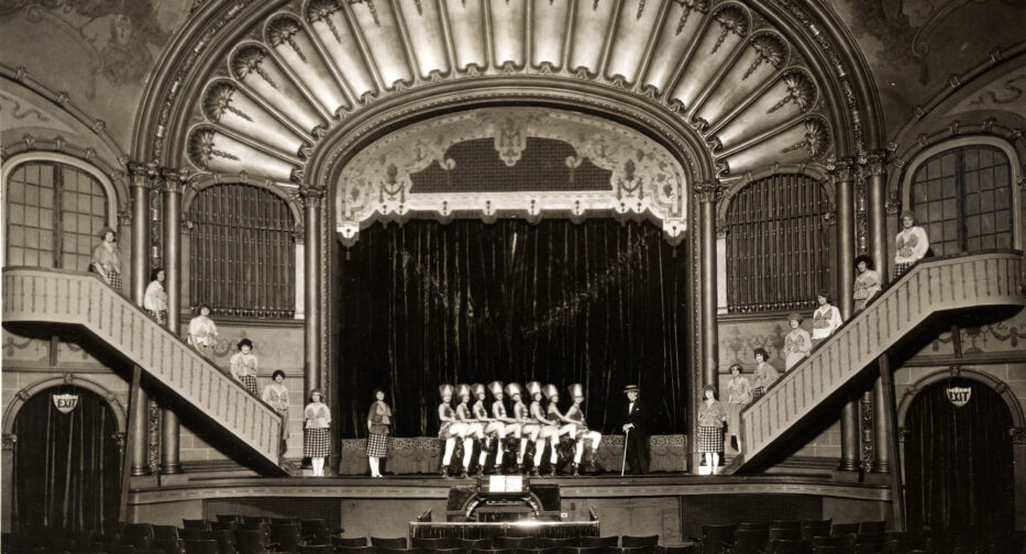 A historic photo of the Liberty Theater showing dancers on stage with additional actors on stairs rising from stage-left and stage-right.