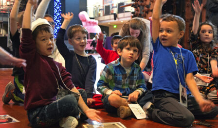 Children sitting on a floor during a field trip at MOHAI and raising their hands to answer questions.