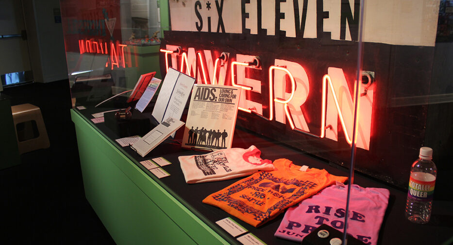 The 611 Tavern sign that was included in the Revealing Queer exhibit. In front of the sign lie an orange shirt and a pink shirt as well as paper paraphernalia.