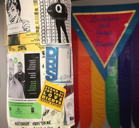 Many one-sheet ads taped to a vertical pole with a pride flag in the background reading, 