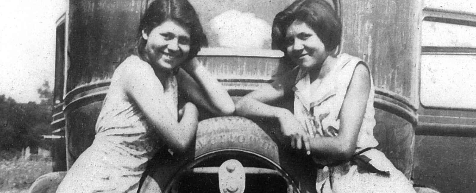 Two girls leaning on back of very old car in black and white.
