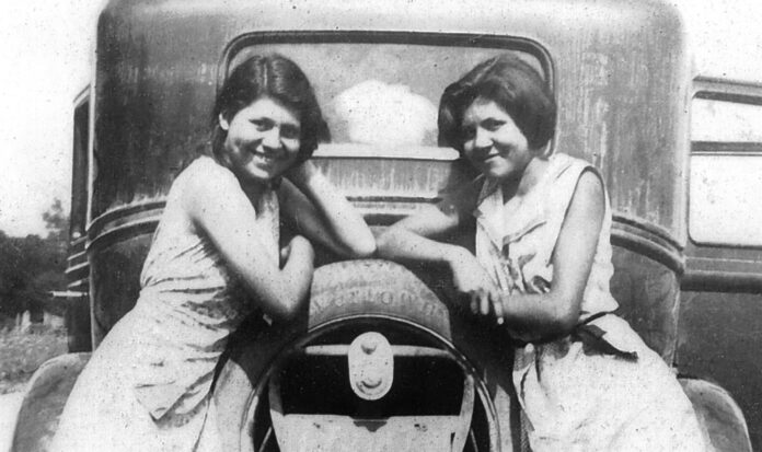 Two girls leaning on back of very old car in black and white.