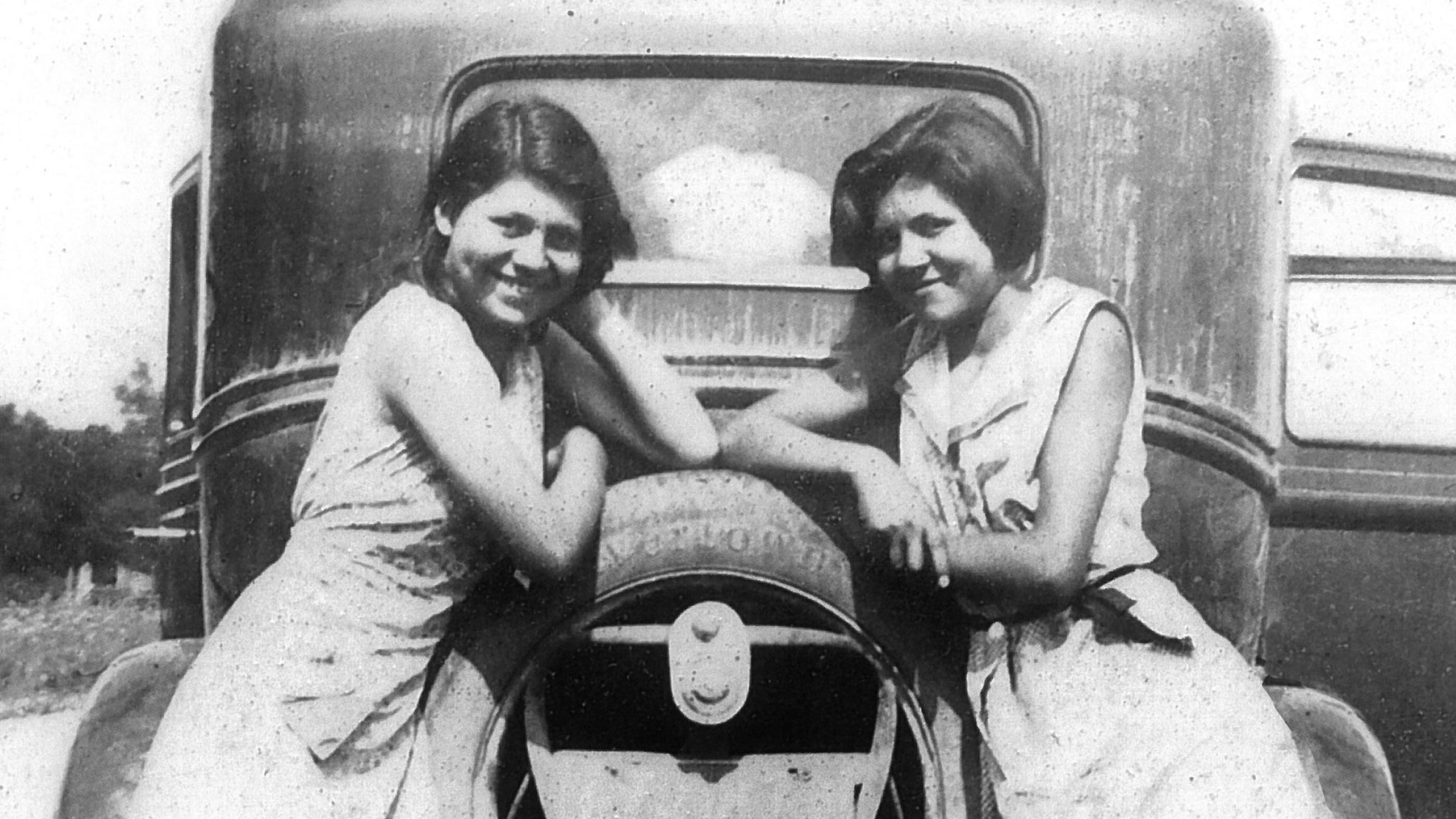 Two girls leaning on back of very old car in black and white.