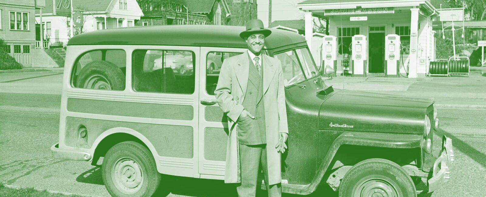 Man in suit with hat standing outside his car. Gas station in the background.