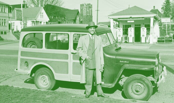 Man in suit with hat standing outside his car. Gas station in the background.
