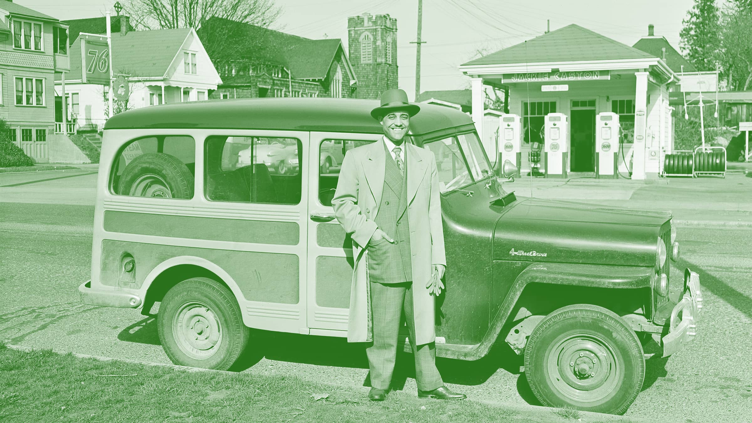 Man in suit with hat standing outside his car. Gas station in the background.