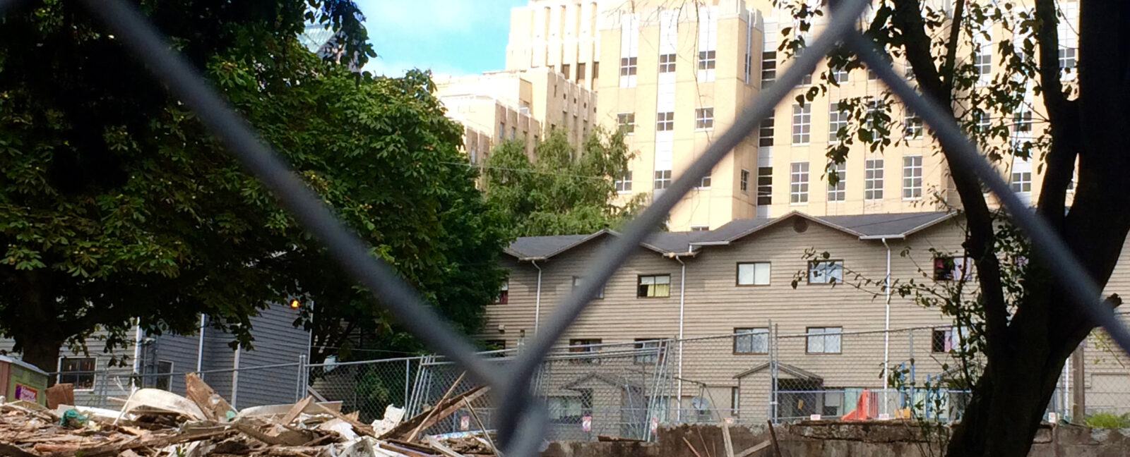 View through chainlink fence of remains of demolished building. Larger buildings in the background.