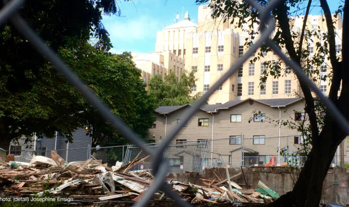 View through chainlink fence of remains of demolished building. Larger buildings in the background.