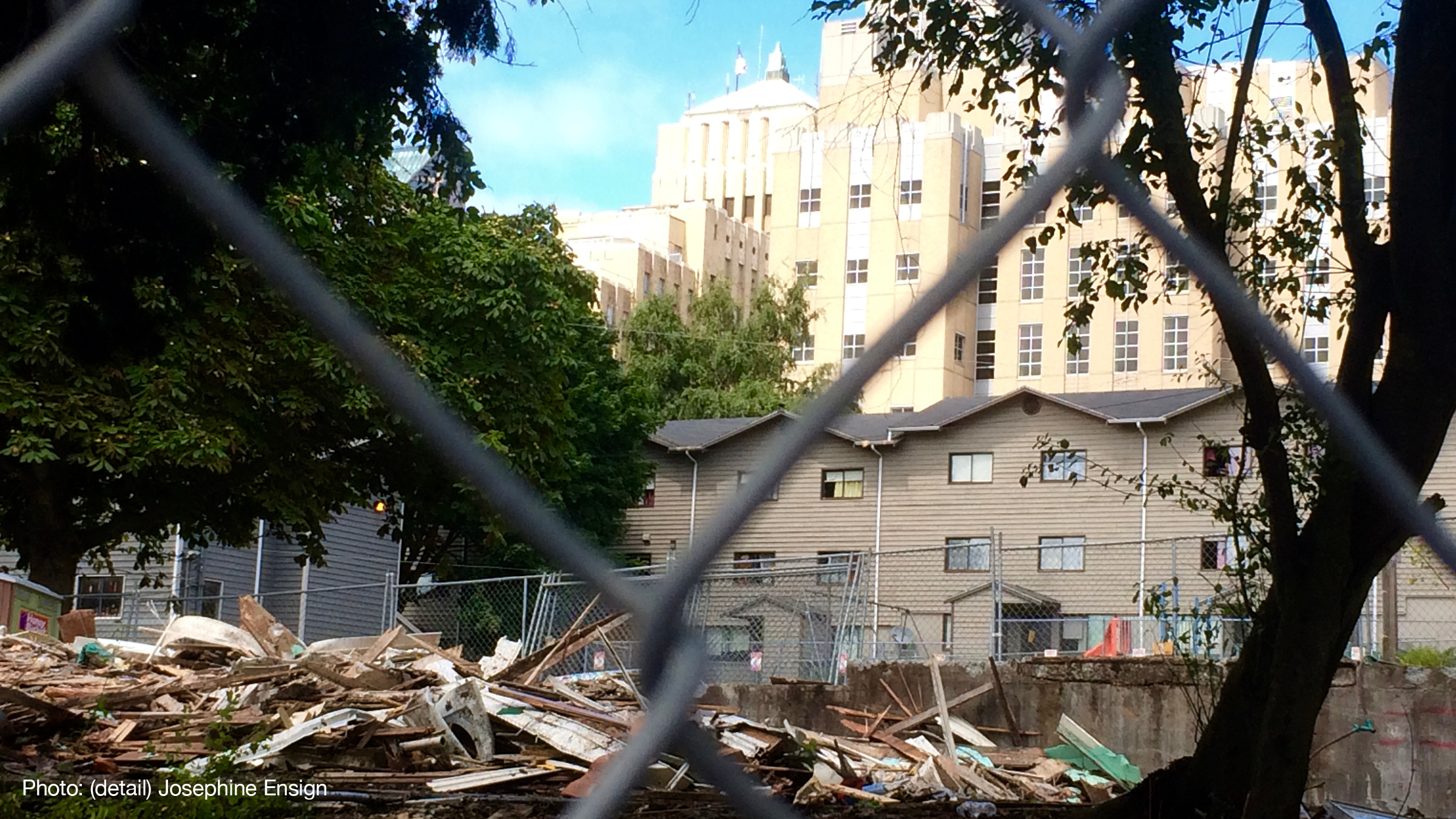 View through chainlink fence of remains of demolished building. Larger buildings in the background.