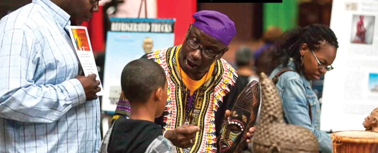 Man in traditional African dress holds traditional wooden mask for visitors to The Unspoken Truths Exhibit.