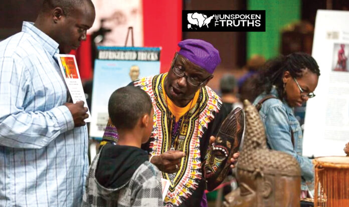 Man in traditional African dress holds traditional wooden mask for visitors to The Unspoken Truths Exhibit.