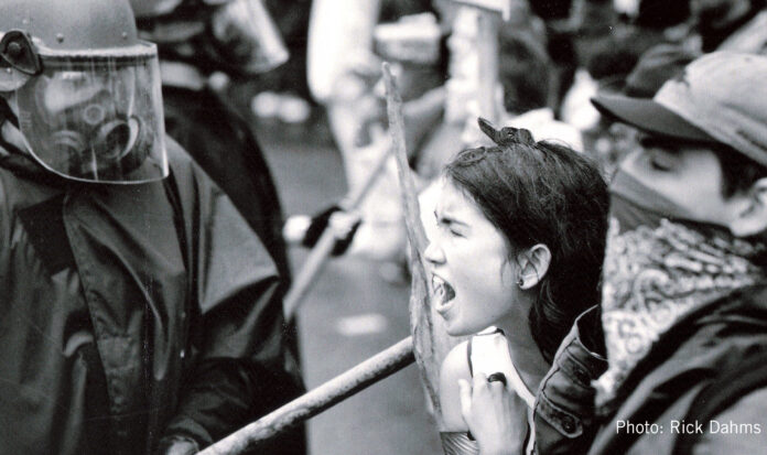 A woman shouting at a person in black combat fatigues wearing a helmet with a clear face shield.