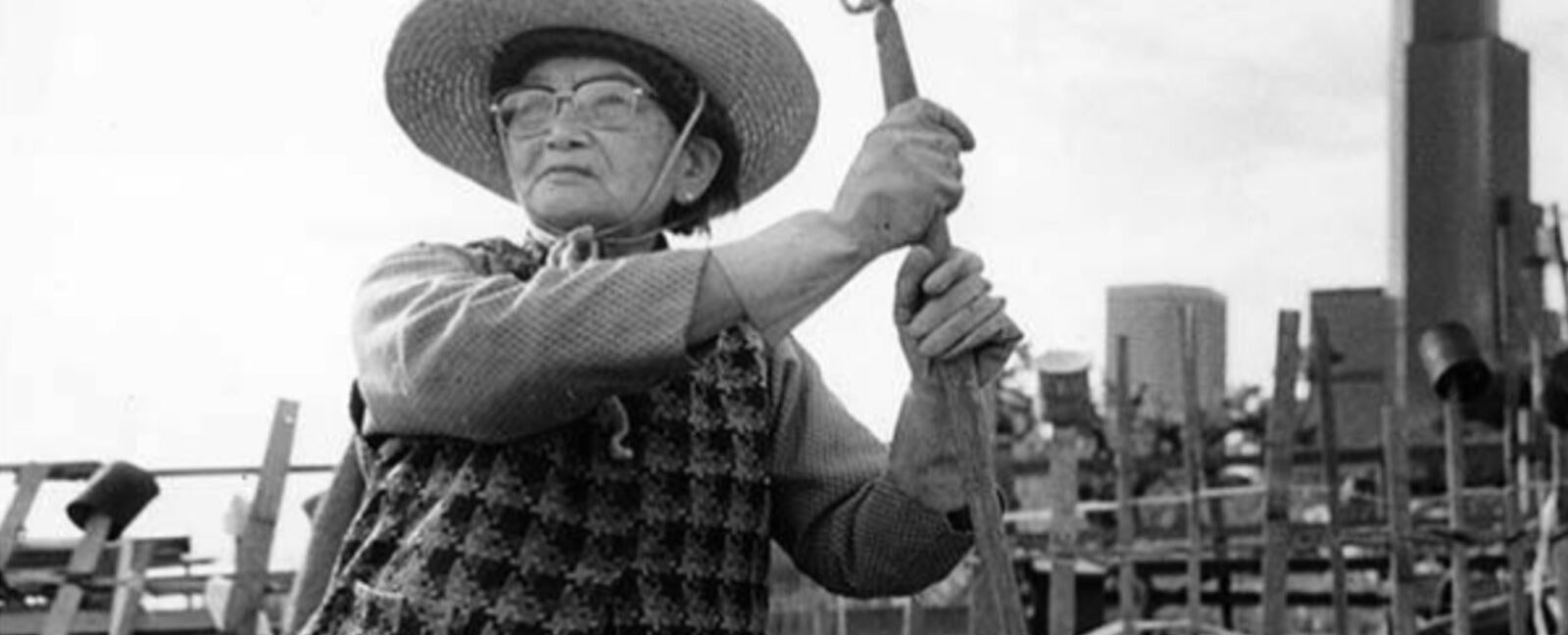 A woman with glasses and a wide brimmed hat stands with a hoe.