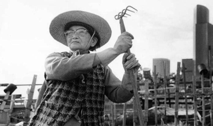 A woman with glasses and a wide brimmed hat stands with a hoe.