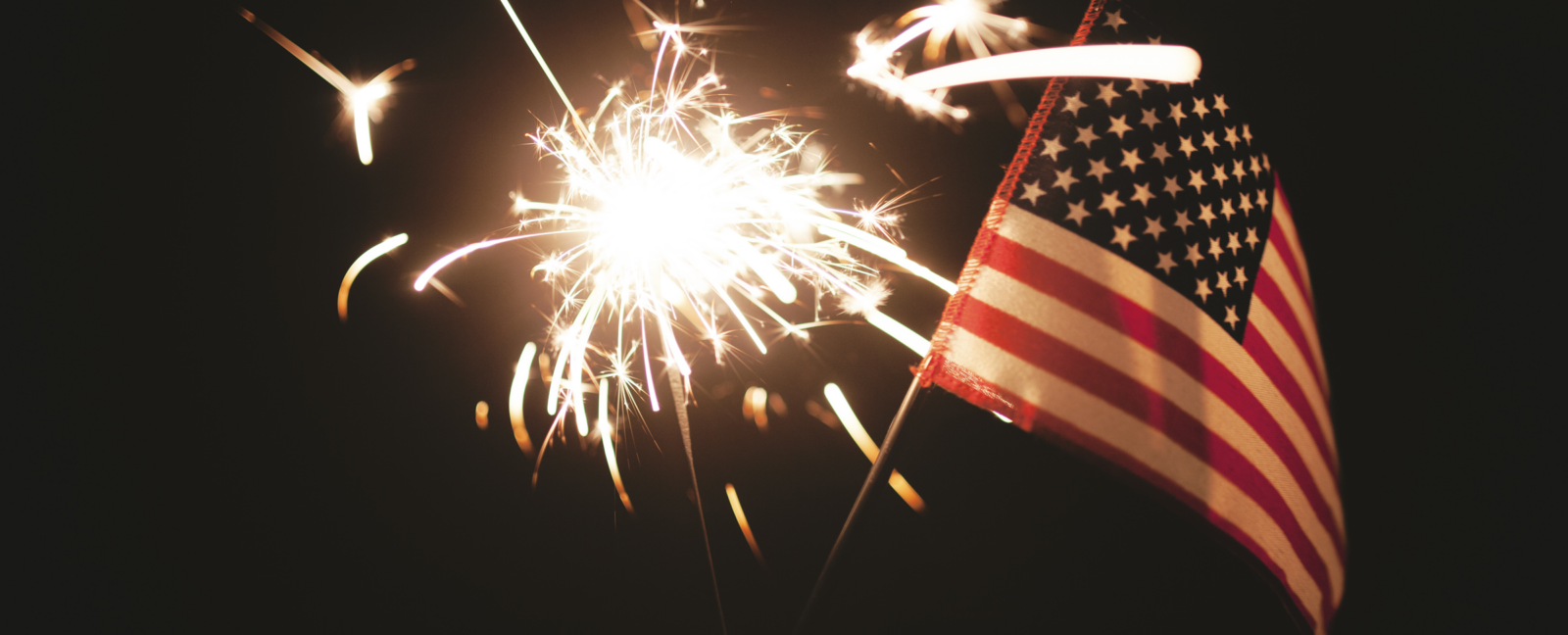 A hand holds a small American Flag and a lit sparkler.