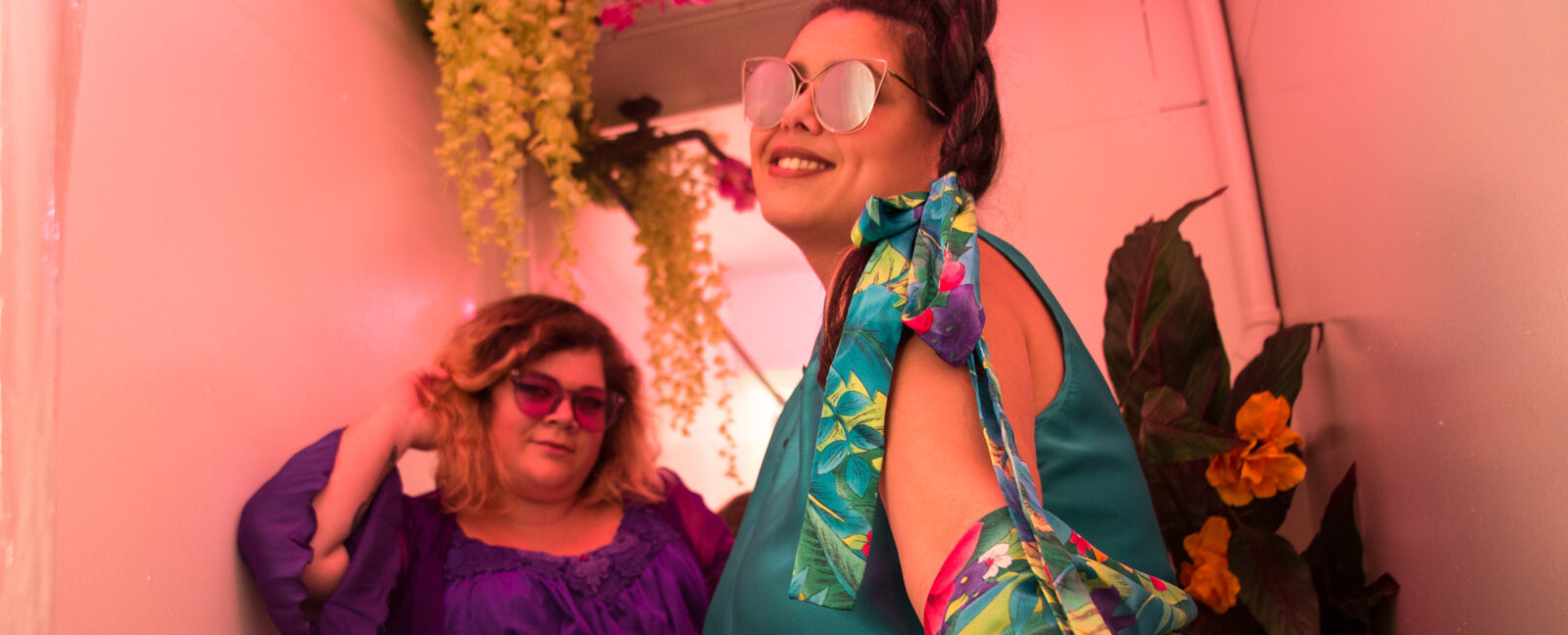 Two women wearing colorful outfits and funky sunglasses smile for the camera. Yellow and pink flowers hang from above.