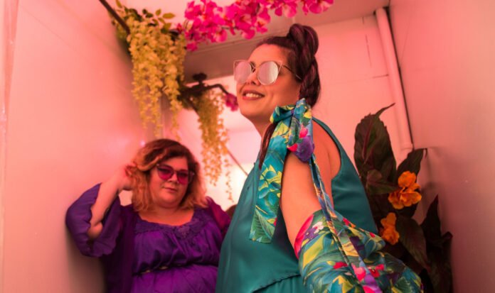 Two women wearing colorful outfits and funky sunglasses smile for the camera. Yellow and pink flowers hang from above.