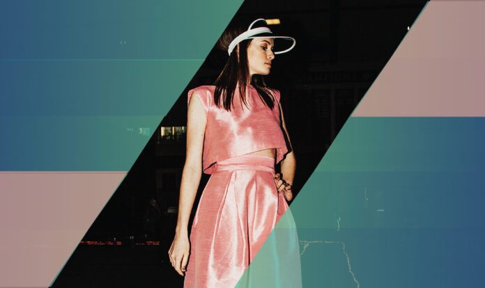 A woman in a pink dress and hat stands against a black background bordered by blue and pink.