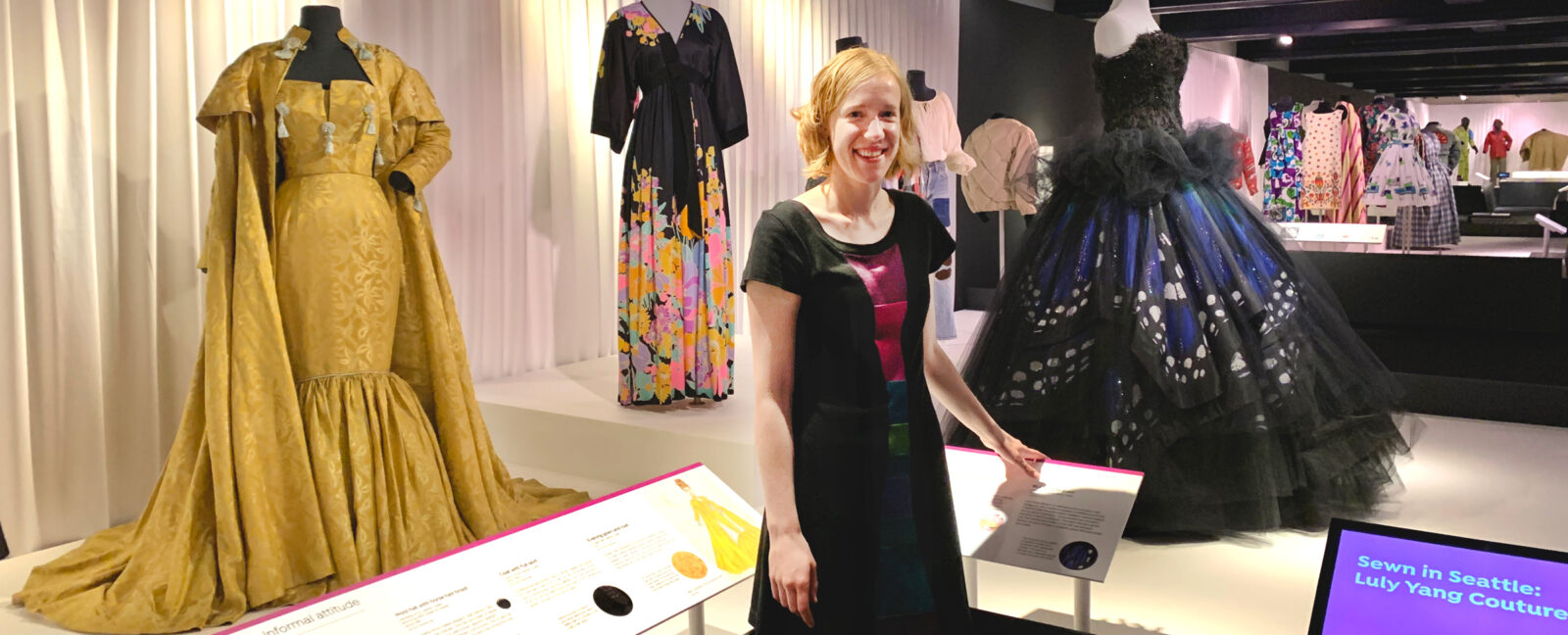 Clara Berg wearing a black and purple dress stands among an exhibit of many other dresses.