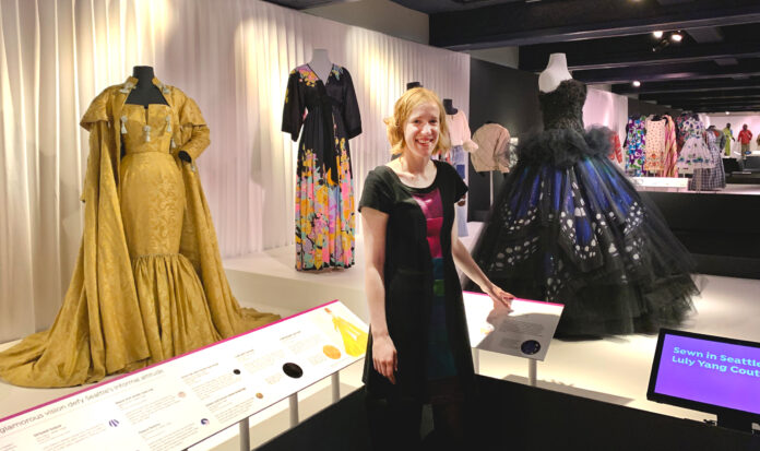 Clara Berg wearing a black and purple dress stands among an exhibit of many other dresses.