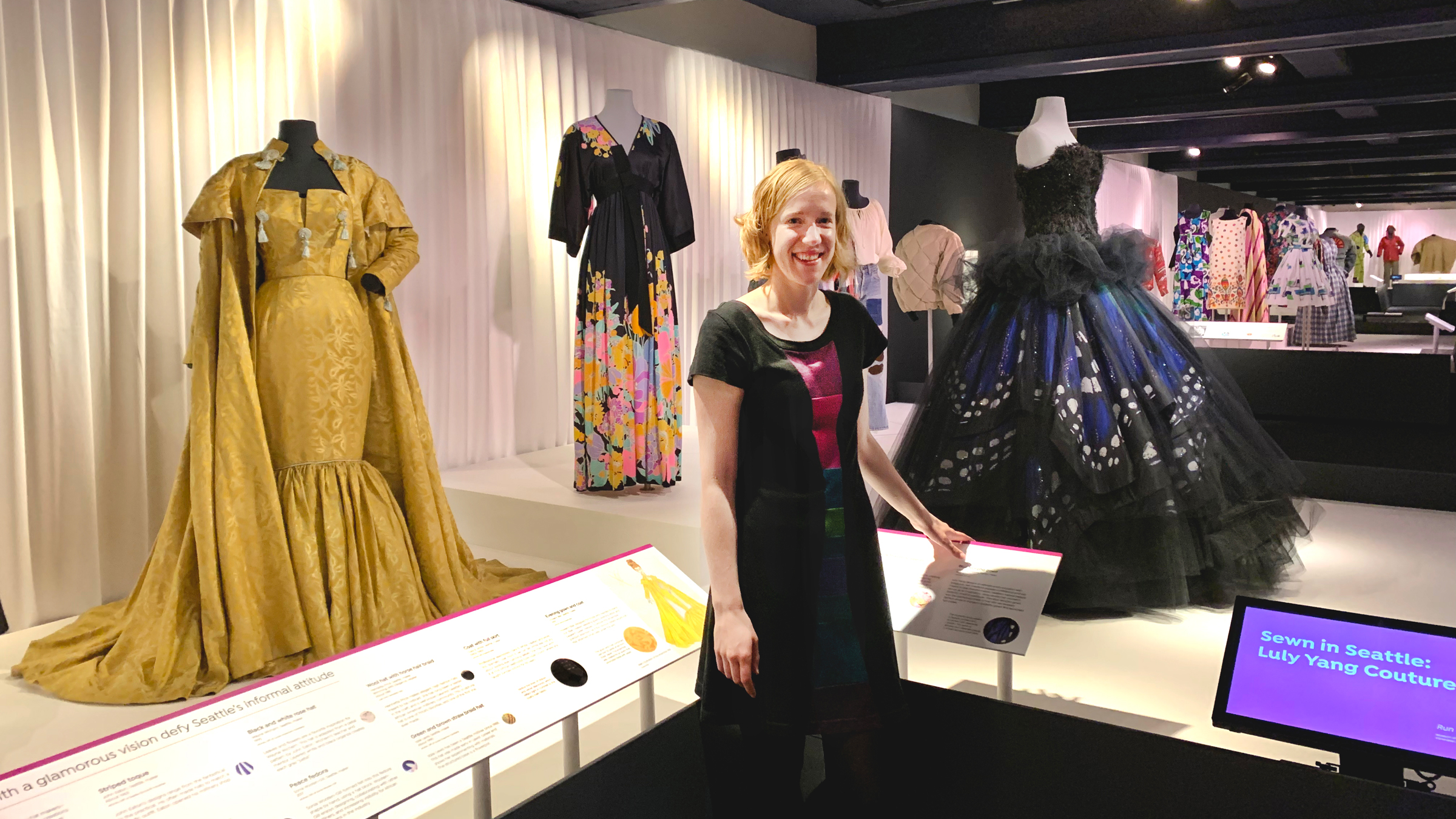 Clara Berg wearing a black and purple dress stands among an exhibit of many other dresses.