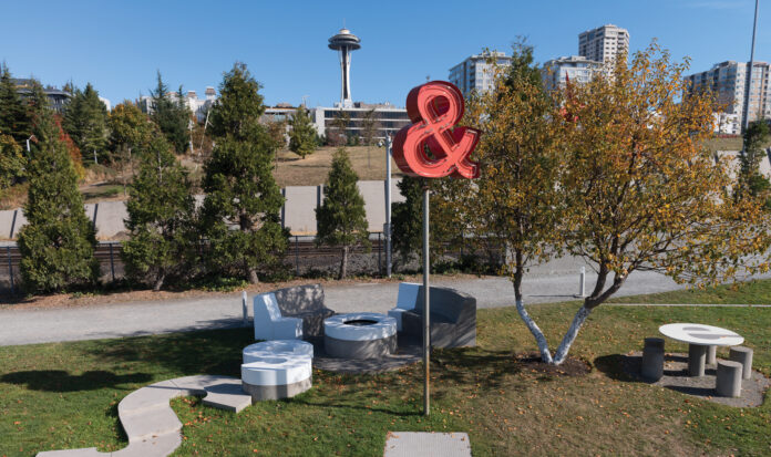 A red ampersand neon sign on a post in downtown Seattle. The Seattle Space Needle can be seen in the background.