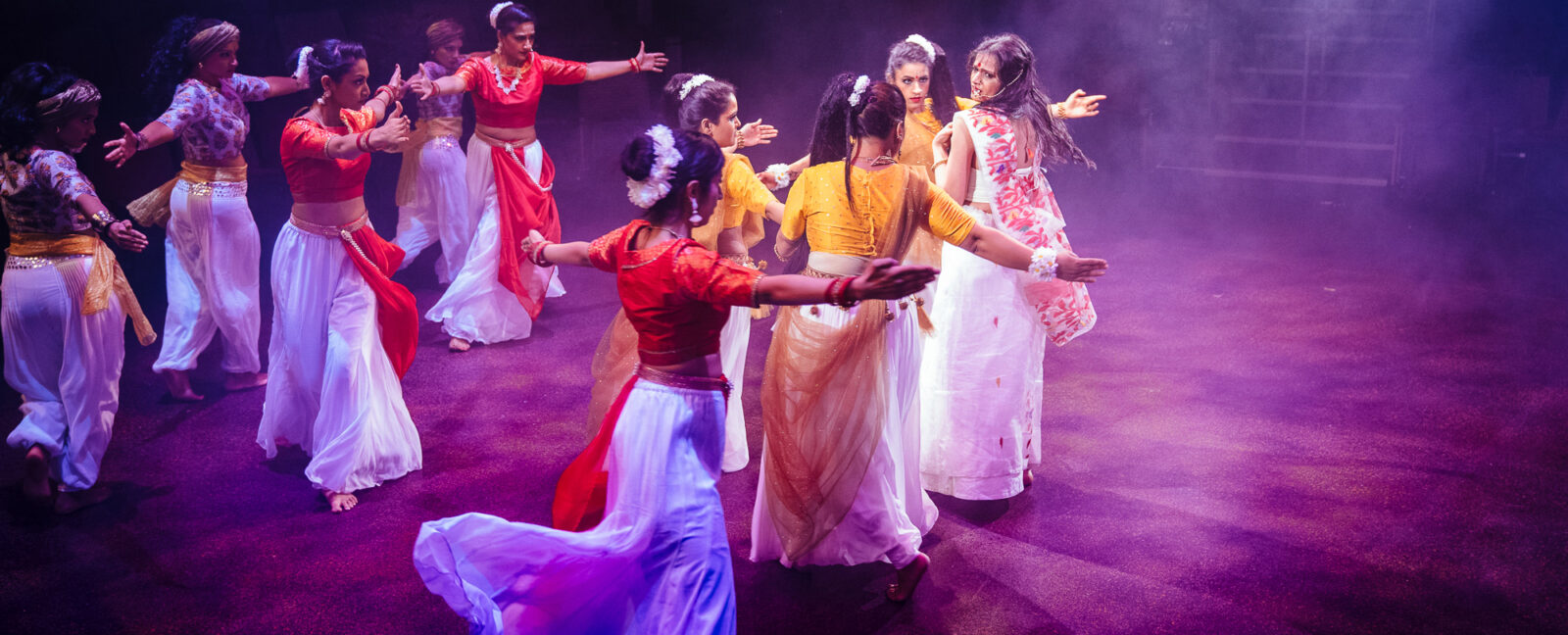 Bollywood dancers wearing white gauchos and colorful tops. The dance floor is lit by pinkish purple light.