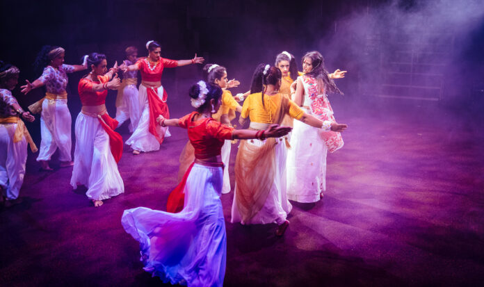 Bollywood dancers wearing white gauchos and colorful tops. The dance floor is lit by pinkish purple light.