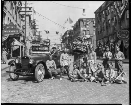 Black and white photograph of a group of dancers posing with a Lion Dance costume and musical instruments next to a car.